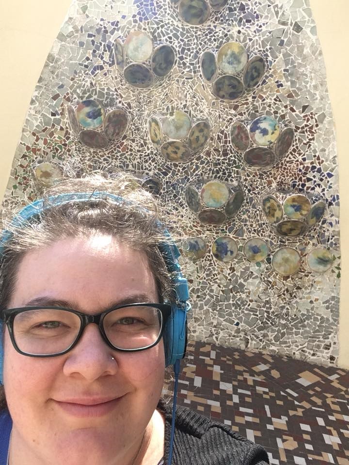 Susie wearing headphone smiling at the camera. Behind her is Gaudi's work in Barcelona. 